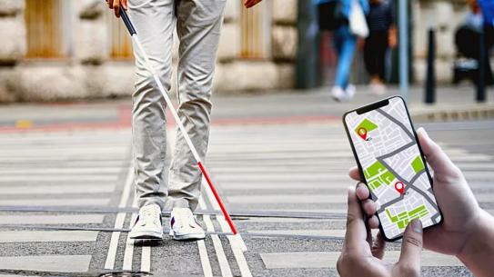 Person walking a crosswalk with a cane and a phone displaying a map application