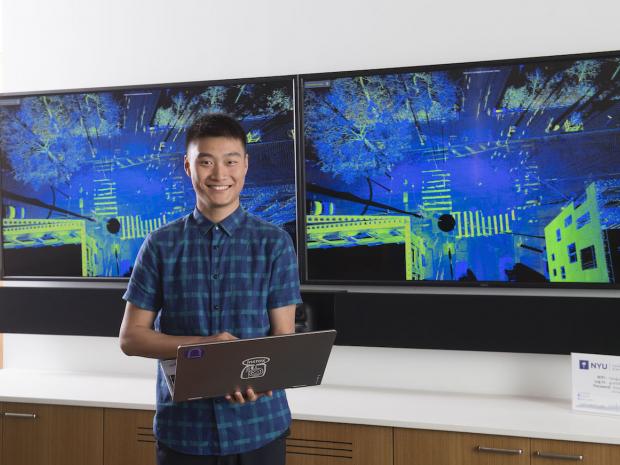 smiling asian male student holding laptop in front of large digital screen