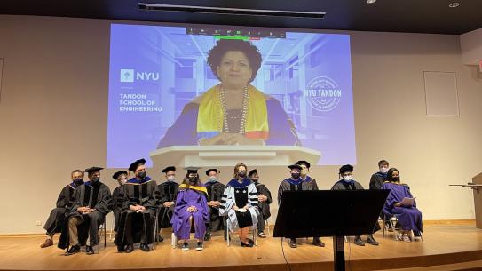 faculty on stage in academic gowns with Chandrika Tandon projected on screen in background
